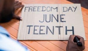 The image shows a person writing "FREEDOM DAY JUNE TEENTH" in bold black letters on a cardboard sign placed on an wooden table.