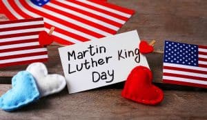 The image shows a card reading Martin Luther King Day surrounded by small American flags and red, white and blue felt hearts on a wooden surface.